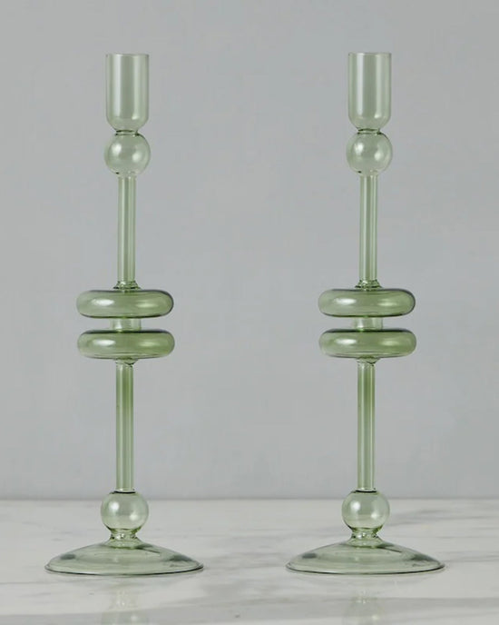 The etúHOME Sage Glass Candlesticks Set of 2 - Large, crafted from recycled glass in translucent green with round and disc accents, stand side by side on a white marble surface.