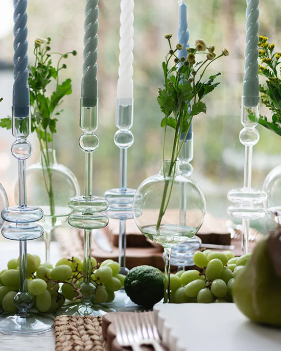 A close-up of a table setting featuring etúHOMEs Denim Glass Candlesticks Set of 2 - Medium, twisted taper candles, small vases with greenery, and bunches of green grapes.