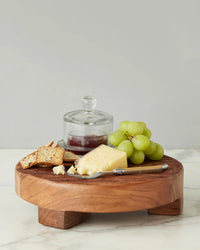 A wooden cheese board with crackers, a wedge of cheese, green grapes, and an etúHOME Bistro Glass Jam Jar, Round filled with jam sits alongside a cheese knife on a marble surface.