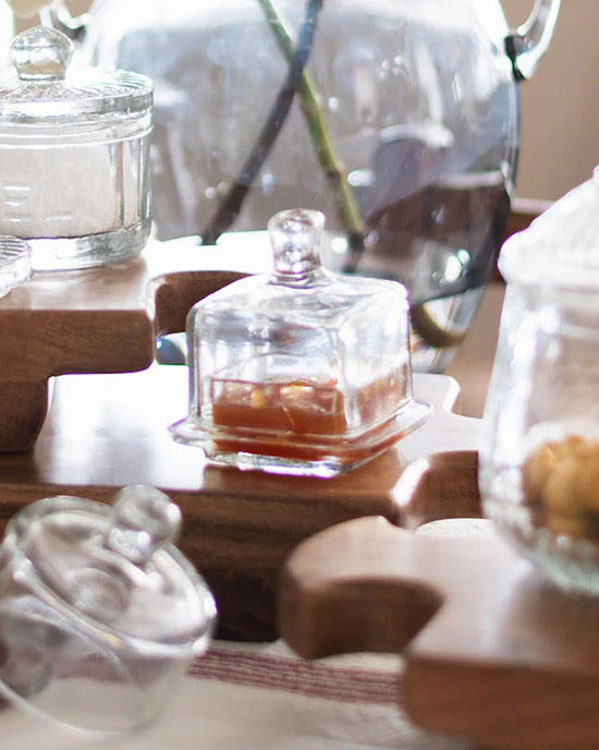 EtúHOME Bistro Glass Jam Jar, Square, by etúHOME, sits on a wooden serving board. Some jars are filled with jam, while a glass vase decorates the background.