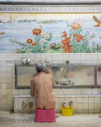 An elderly person with gray hair sits on a pink stool at the tiled Hot Springs bathhouse by Random House, surrounded by bright floral and bird murals above, with nearby buckets and taps completing the traditional charm.