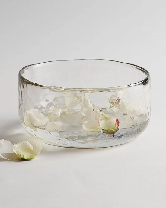The Collective Pebbled Salad Bowl in clear recycled glass holds water with floating white flower petals, while a few petals and a bud rest on the white surface beside it.