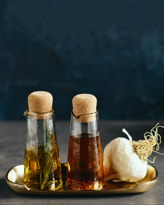 The Collective Pebbled Small Carafe Set features two hand-blown glass carafes with cork stoppers on a gold tray—one for olive oil and herbs, the other for vinegar—styled alongside a garlic bulb.