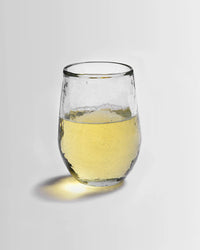 A Pebbled Small Glass from The Collective, filled halfway with pale yellow liquid, rests on a plain white surface and casts a slight shadow to the left.