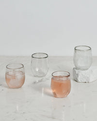 The Collective Pebbled Spirits Glasses, set of 4, are shown on a white marble surface—with two glasses filled with a pink drink and ice, and two empty; one glass is placed on a white stone.