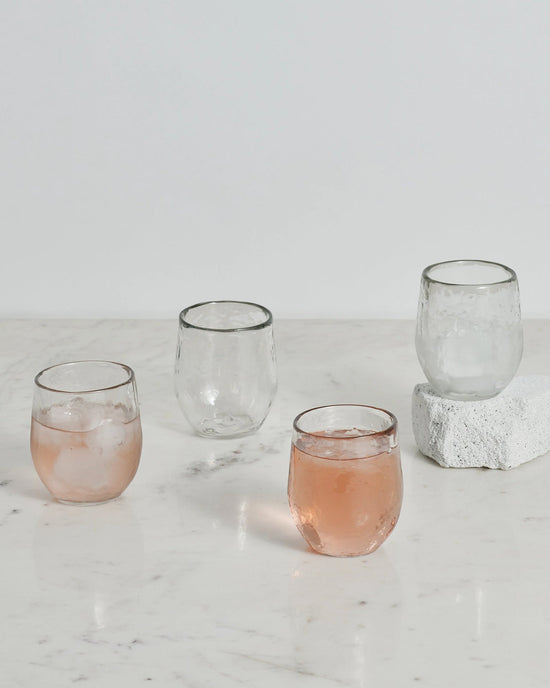 The Collective Pebbled Spirits Glasses, set of 4, are shown on a white marble surface—with two glasses filled with a pink drink and ice, and two empty; one glass is placed on a white stone.