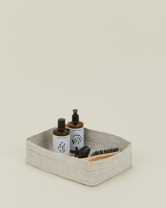 The Hawkins New York Essential Woven Storage - Letter Tray in beige holds two minimal pump bottles and a black hairbrush, all set against a simple light background.