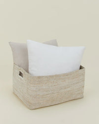 The Essential Woven Storage - Low Basket by Hawkins New York, featuring two handles, holds two large pillows—one white, one light beige—against a simple background, making it perfect for stylish blanket or pillow storage.