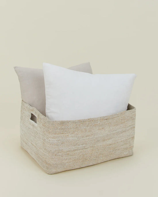 The Essential Woven Storage - Low Basket by Hawkins New York, featuring two handles, holds two large pillows—one white, one light beige—against a simple background, making it perfect for stylish blanket or pillow storage.