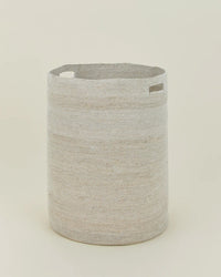 The Essential Woven Hamper by Hawkins New York is a round, light beige handwoven seagrass hamper with two rectangular cutout handles, shown upright against a plain off-white background.