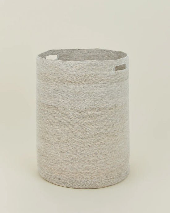 The Essential Woven Hamper by Hawkins New York is a round, light beige handwoven seagrass hamper with two rectangular cutout handles, shown upright against a plain off-white background.