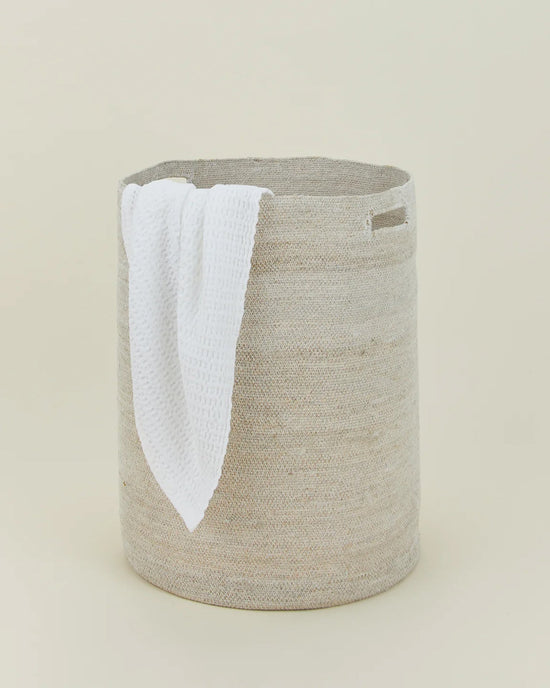 The Hawkins New York Essential Woven Hamper, a round beige handwoven seagrass basket with a rectangular handle cutout, holds a white textured towel draped over its side against a plain light background.