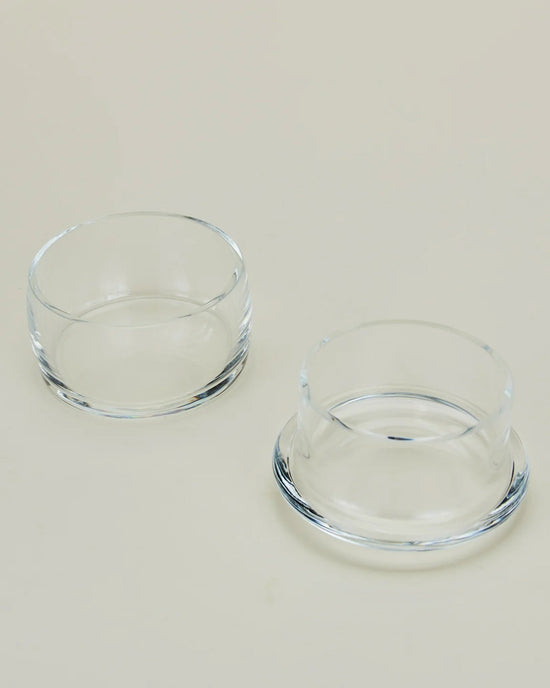 Two small, clear handblown glass bowls from Hawkins New Yorks Simple Glass Butter Keeper are placed on a light beige surface—one upright, one upside down—highlighting their smooth, minimalist design.
