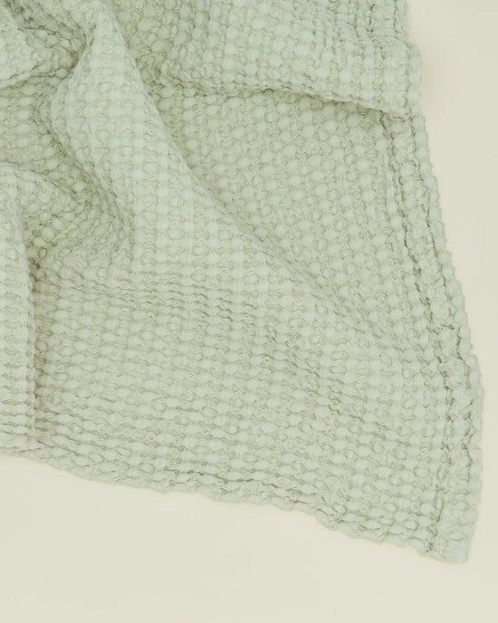 A close-up of the Hawkins New York Simple Waffle Bath Sheet in Sage, featuring a textured, light green waffle weave pattern, shown slightly folded on a light surface.
