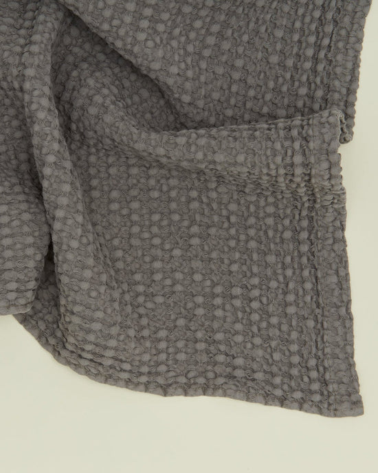A close-up of the Hawkins New York Simple Waffle Bath Towel in Dark Grey shows its textured, waffle weave and folded corner, highlighting detailed stitching—crafted from soft, stonewashed cotton.