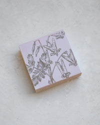 A square, pale pink box labeled "Hudson Valley Matches | Bleeding Hearts" by The Floral Society sits on a textured light surface. Its lid features hand-drawn wildflowers and botanical line art, with The Floral Society along the edge.