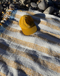 A yellow baseball cap rests on the caminito Suave Miel - Blanket Roll, handwoven in Mexico with white and yellow stripes, spread over rocky ground with smooth gray stones around the edges.