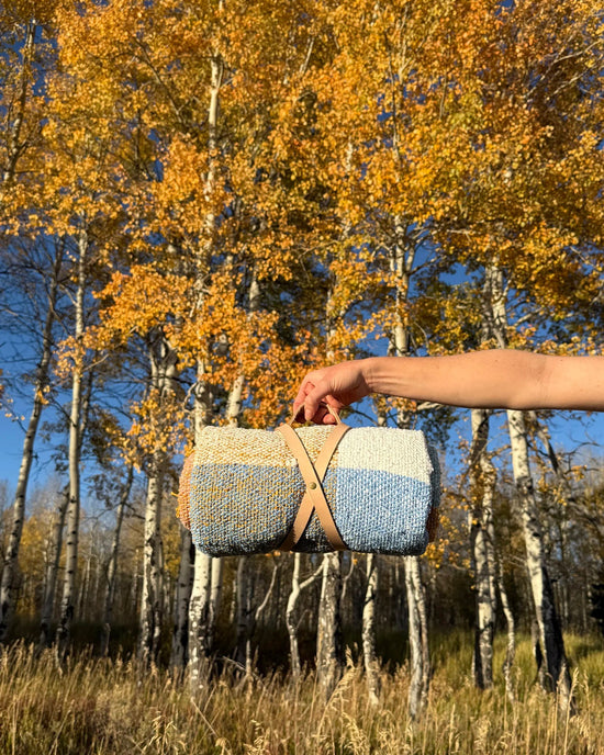 A hand holds the Suave Campo - Blanket Roll by caminito, featuring a rolled-up, multicolored design with a tan leather strap, set against tall autumn trees under a clear blue sky.