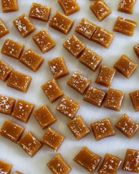 Wildflower Caramel Co Maple Whiskey Caramels in a Bag are neatly arranged on a white surface, each topped with sea salt.