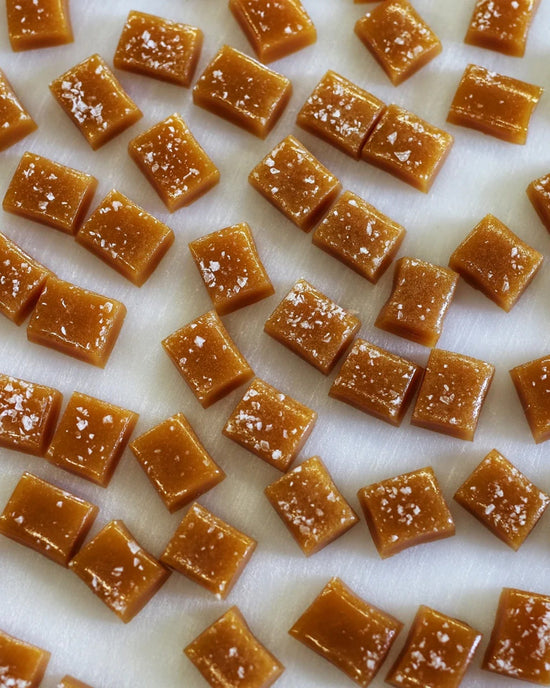 Wildflower Caramel Co Maple Whiskey Caramels in a Bag are neatly arranged on a white surface, each topped with sea salt.