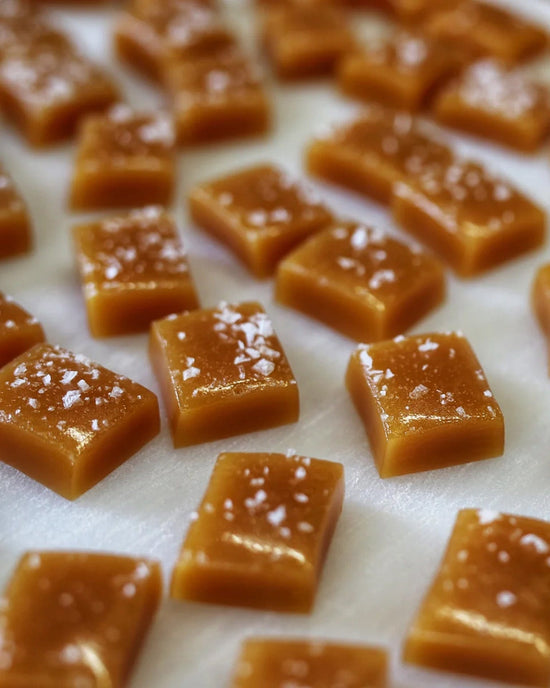 Wildflower Caramel Co’s Coconut Caramels (Vegan) in a Bag are arranged on a white surface, with each rectangular piece topped with coarse sea salt.