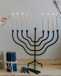 A hand lights the last Hanukkah candle on the Beacon Menorah - Tall by The Floral Society, with all candles aglow. The wrought iron menorah sits on a wooden surface next to bundles of white and blue candles.