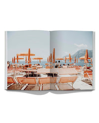 An open book, "Italian Summer" by Simon & Schuster, displays a photo of Italy's coast with orange lounge chairs, umbrellas, people relaxing, and distant mountains under a clear sky.