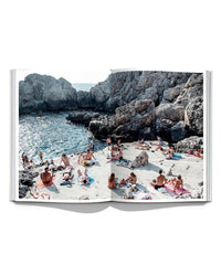 Photo of the "Italian Summer" book by Simon & Schuster, open to a page featuring a beach scene inspired by Italian coastal towns, with sunbathers on towels beside rocky cliffs and sea—evoking Natalie Obradovich’s photographic style.