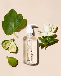 A clear pump bottle of Rosie Jane James Everyday Body Wash sits on a beige background, surrounded by fig leaves, green figs, and a white flower—a fresh scene for this hydrating creamy cleanser.