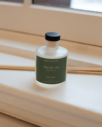 The Near & Native Room Diffuser in Fraser Fir, featuring a black cap and a green label with white text, sits on the windowsill with several wooden reeds, bringing a fresh touch to the space.