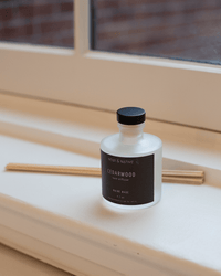 A frosted glass bottle labeled Near & Native Room Diffuser in Cedarwood with a black cap sits on a windowsill beside several reed sticks, with a window and brick wall providing the backdrop for this refreshing cedarwood scent.