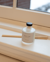 A Near & Native Room Diffuser in White Pine with a black cap rests on a windowsill beside several natural-colored reed sticks.