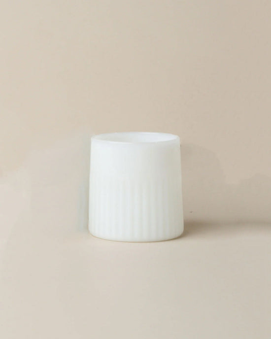 The Kairos Medium Water Glass in Opaque White by Saarde, featuring vertical grooves and a cylindrical shape, rests on a beige surface with a matching background.