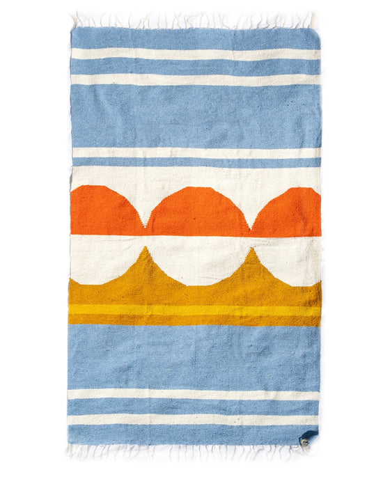 The Luna Paseo - Blanket Roll by caminito is a rectangular woven rug with blue and white stripes, orange semicircles in the center, white and yellow bands, fringed edges, is lightweight, machine washable, and perfect for outdoor use.