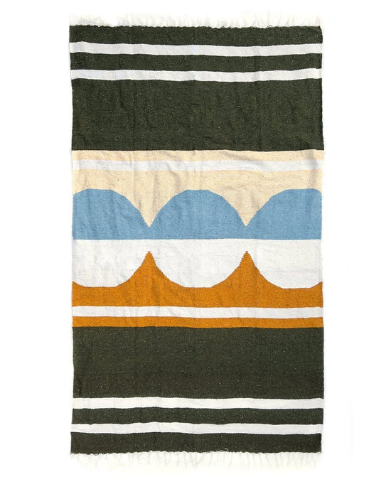 The Luna Lago - Blanket Roll by caminito is a woven, lightweight blanket featuring black, white, tan, yellow, and blue stripes with abstract blue and orange arches at the center and fringed short edges—ideal for outdoor use.