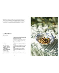 A bowl with blue floral patterns filled with roasted olives sits outdoors on a green-checkered cloth, recipe for Olive Calde at left. Scene inspired by traditional Italian fare from "La Nostra Cucina Povera – Everyday Italian Eats" by Cozy Publishing.