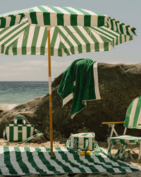 The Business & Pleasure Co. Amalfi Umbrella - Crew Canyon Green Stripe, plus towel, bag, chair, and mat are set up on a sandy beach with rocks nearby and the ocean gleaming in the background on a sunny day.