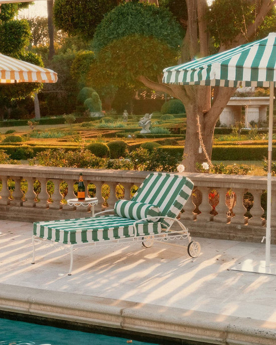 A Bolster Pillow - Crew Canyon Green Stripe by Business & Pleasure Co rests on a striped lounge chair by the pool, shaded by a matching umbrella. A side table holds two glasses and champagne amid manicured gardens, creating a relaxing scene.