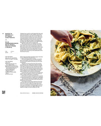 A hand lifts a square ravioli topped with pesto from a bowl, set on a white lace tablecloth beside Chronicle Books’ "Pasta Grannies: Comfort Cookbook"—a taste of authentic Italian cuisine.