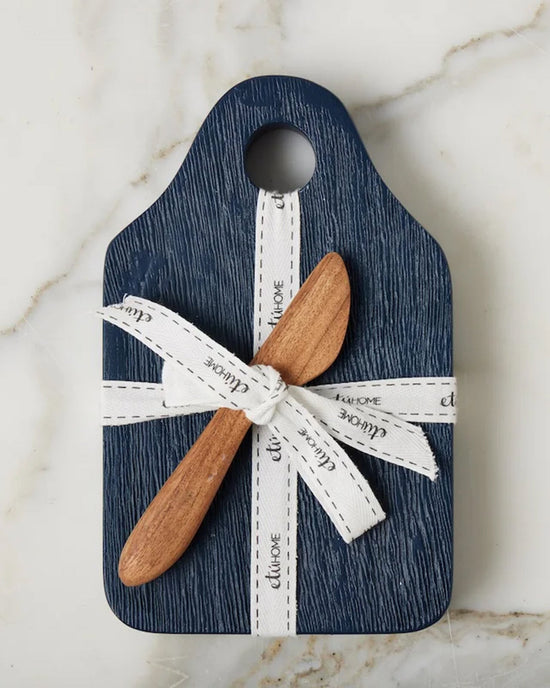 The etúHOME Dutch Small Cheese Board w/ Spreader in Navy Blue features a top hole with a white ribbon and includes a wooden spreader, all elegantly displayed on a marble surface.