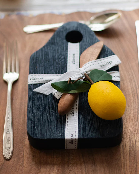 The etúHOME Dutch Small Cheese Board w/ Spreader in Navy Blue, tied with a white ribbon and topped with wooden utensils, green leaves, and a yellow lemon, sits next to a fork and knife on a wooden surface.