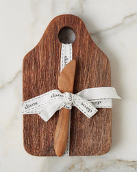 The etúHOME Dutch Small Cheese Board w/ Spreader in Natural features a handy top hole, is tied with a white ribbon, and comes with a wooden spreader—an ideal hostess gift, beautifully presented on light marble.