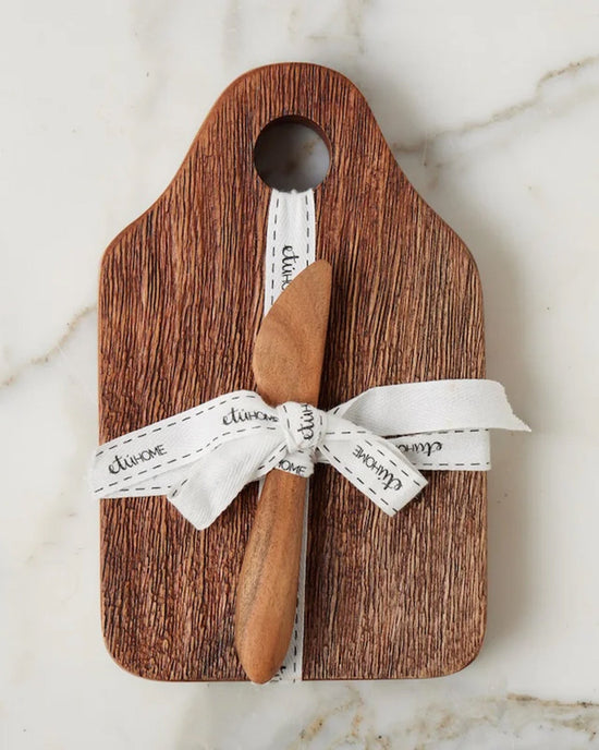 The etúHOME Dutch Small Cheese Board w/ Spreader in Natural features a handy top hole, is tied with a white ribbon, and comes with a wooden spreader—an ideal hostess gift, beautifully presented on light marble.