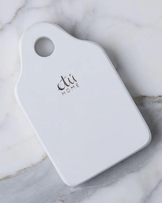 The etúHOME Dutch Small Cheese Board w/ Spreader in White is a small, rectangular ceramic serving board with rounded corners, a handle hole, and etú HOME printed on it, styled after classic Dutch cheese boards.