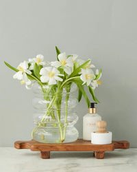 A clear glass vase with white flowers, a lotion bottle, and a small container with a wooden brush rest on the etúHOME Normandy Wood Riser Footed Tray, Medium, atop a marble surface.