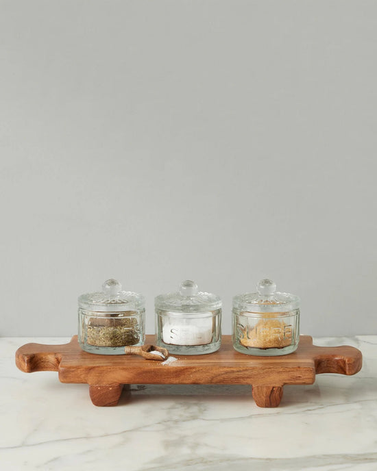 Three glass jars with lids, filled with assorted spices, are displayed on the medium Normandy Wood Riser Footed Tray by etúHOME, placed on marble against a plain gray background.