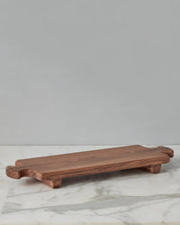 The etúHOME Normandy Wood Riser Footed Tray, Large, features a rectangular Acacia wood design with short legs and handles at each end, shown on a marble surface against a plain gray background.