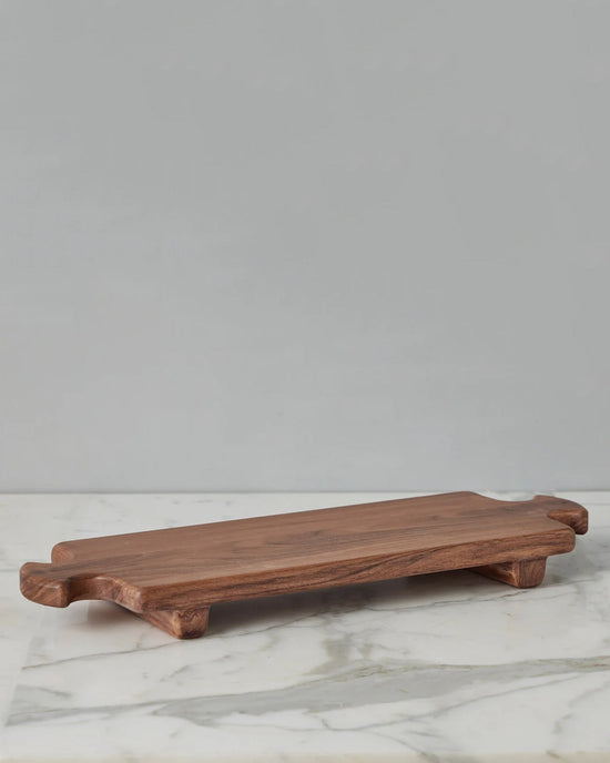 The etúHOME Normandy Wood Riser Footed Tray, Large, features a rectangular Acacia wood design with short legs and handles at each end, shown on a marble surface against a plain gray background.