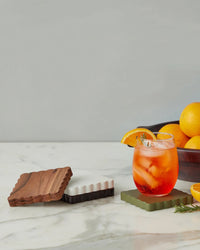 A glass of iced orange cocktail sits on a green coaster beside stacked etúHOME Scalloped Wood Coasters S/4 in Vineyard Colors; a bowl of oranges is in the background on a marble surface.