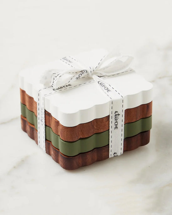 The etúHOME Scalloped Wood Coasters S/4 in Vineyard Colors—white, green, and brown—are crafted from responsibly sourced wood for an eco-friendly touch, tied with a white ribbon and displayed on a marble surface.